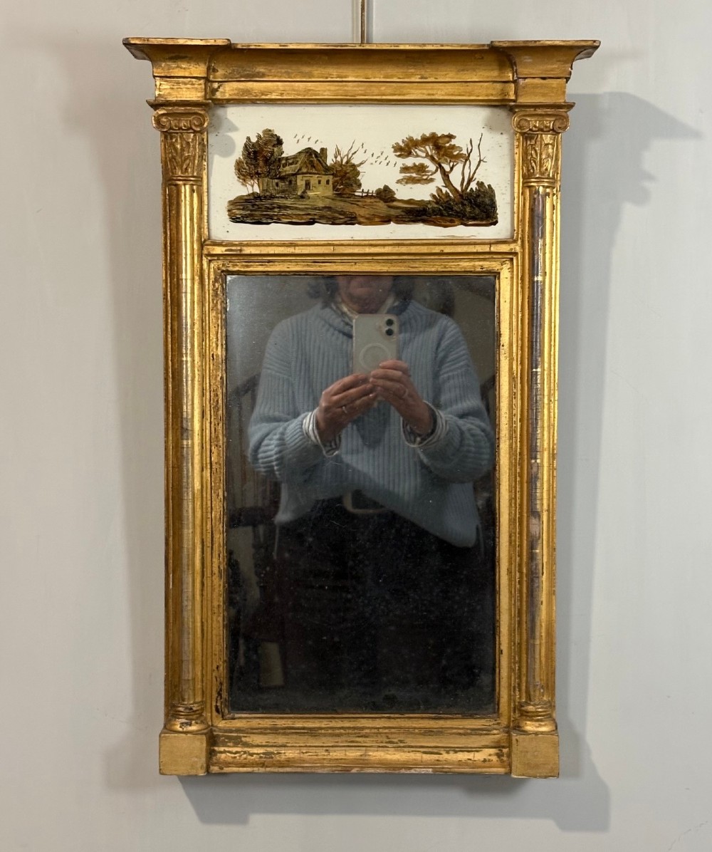 A Regency period giltwood pier glass mirror featuring fluted side columns, a stepped pediment cornice, and an upper verre eglomise glass panel depicting a rural building and landscape.