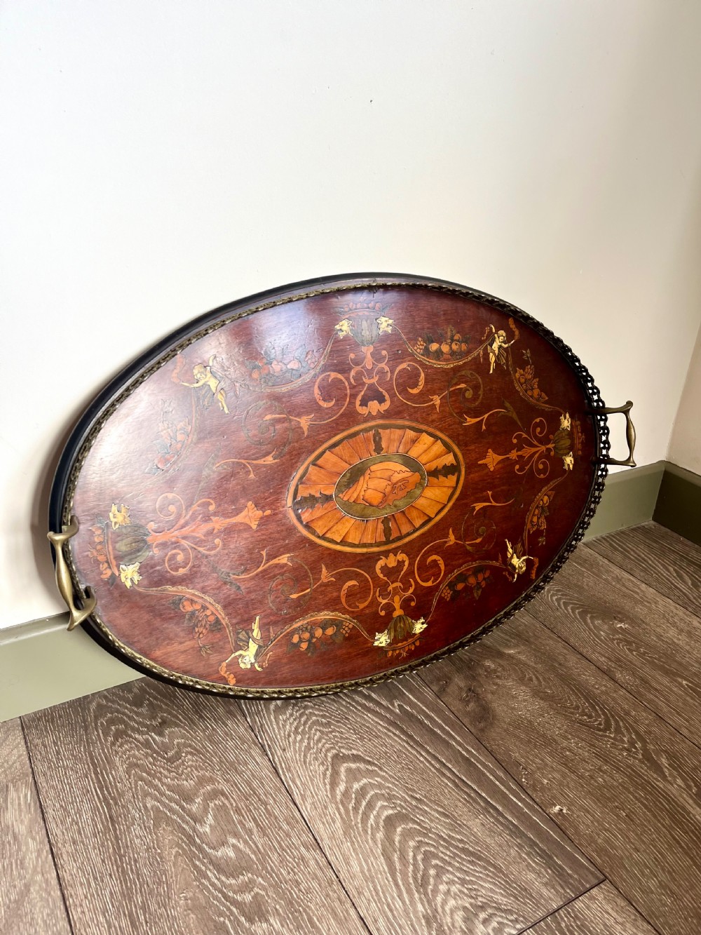 An oval wooden Edwardian serving tray featuring detailed marquetry inlay of scrollwork and cherubs with two brass side handles and a metal rim.