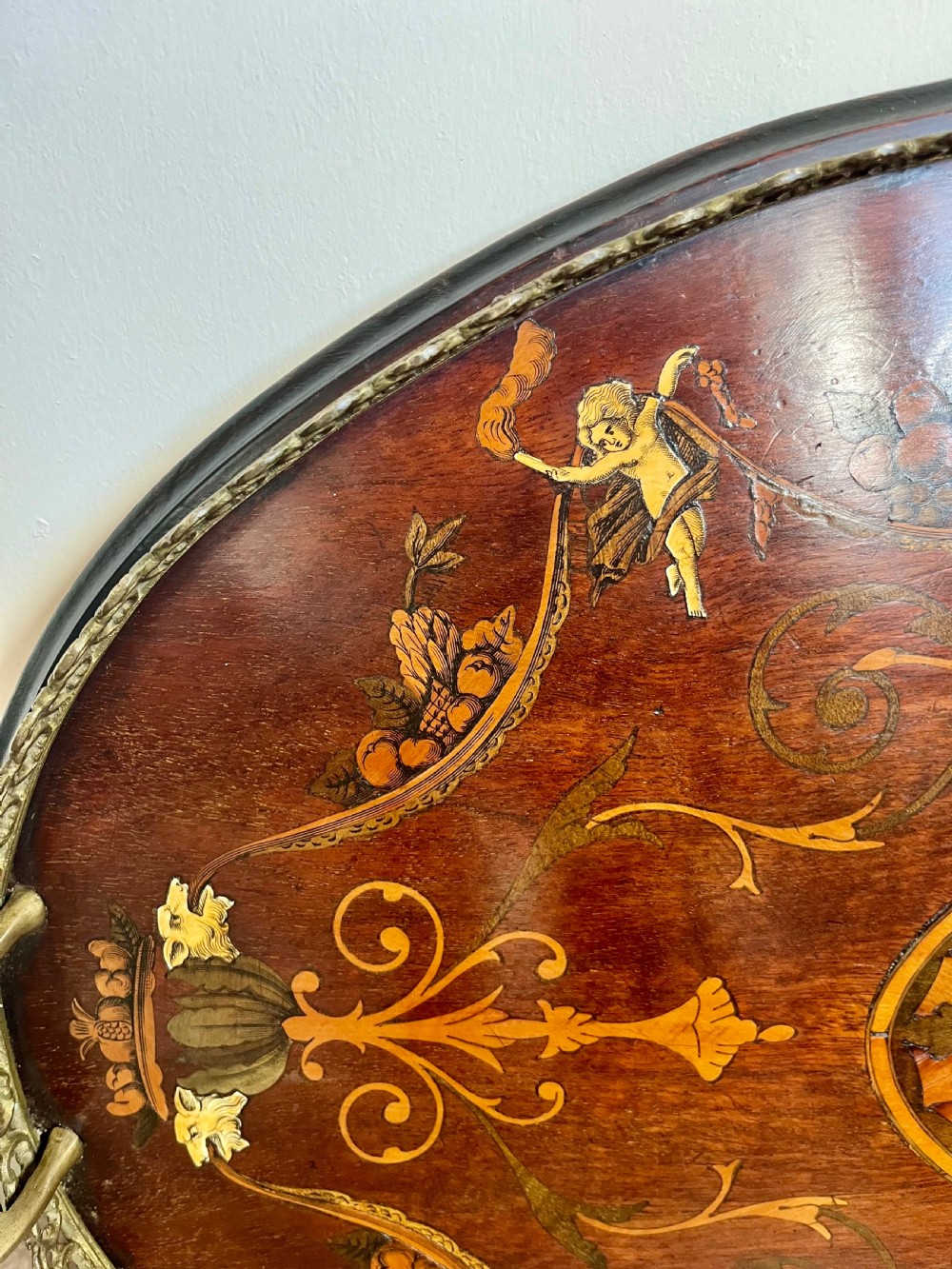An oval wooden Edwardian serving tray featuring detailed marquetry inlay of scrollwork and cherubs with two brass side handles and a metal rim. View III