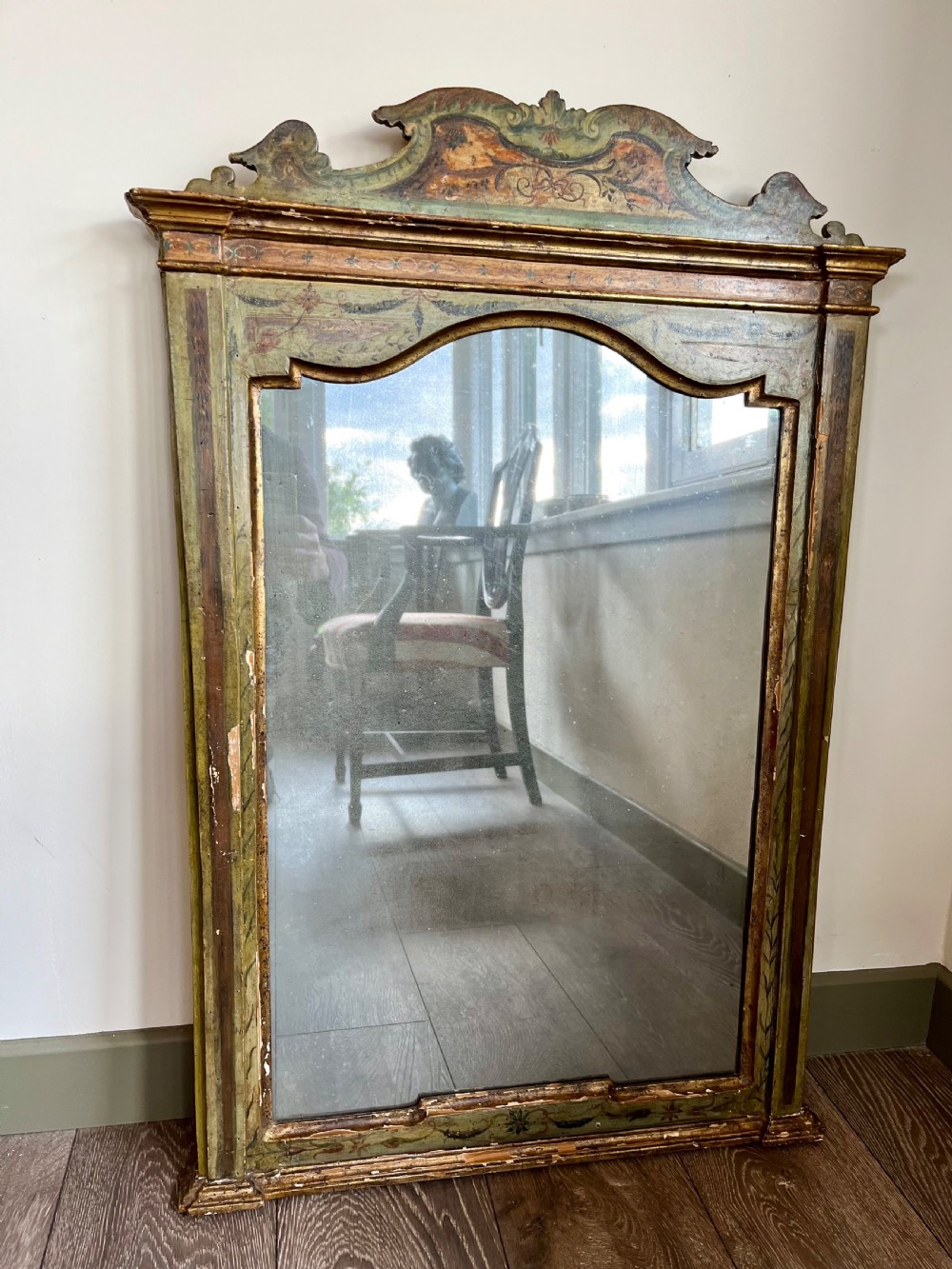 A rectangular painted wooden wall mirror featuring a carved scrolled pediment, an arched inner frame, and painted foliage motifs on a green background.