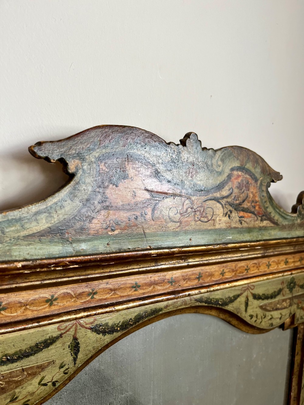 A rectangular painted wooden wall mirror featuring a carved scrolled pediment, an arched inner frame, and painted foliage motifs on a green background. View III