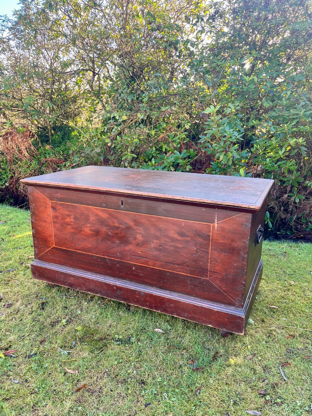 A rectangular dark reddish-brown pine storage chest with a hinged lid, painted panel border detail, and iron side handles.
