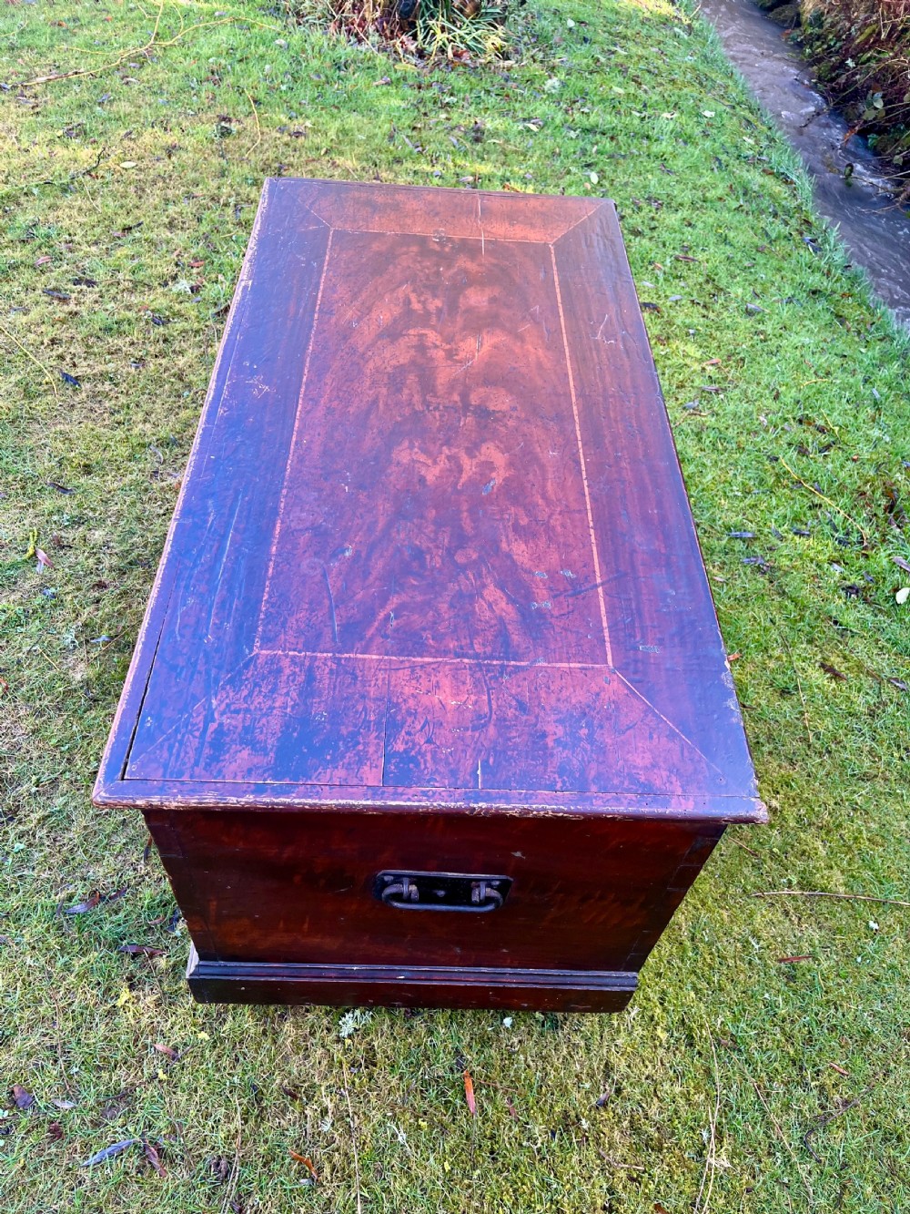 A rectangular dark reddish-brown pine storage chest with a hinged lid, painted panel border detail, and iron side handles. View II