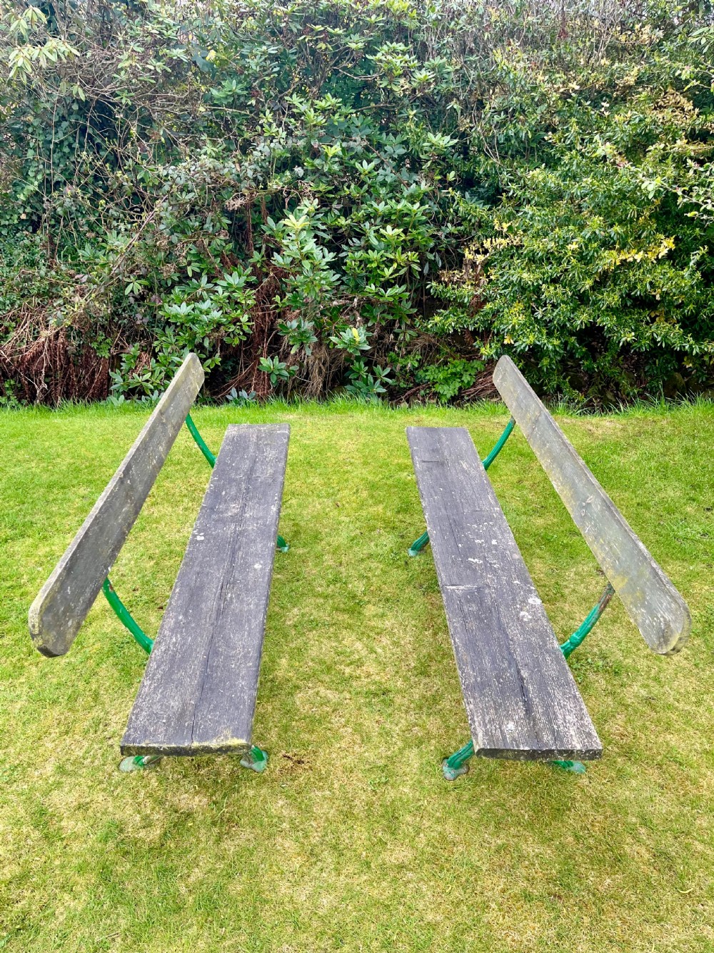 A pair of outdoor garden benches featuring green painted cast iron legs supporting weathered wooden plank seats and backrests. View VIII