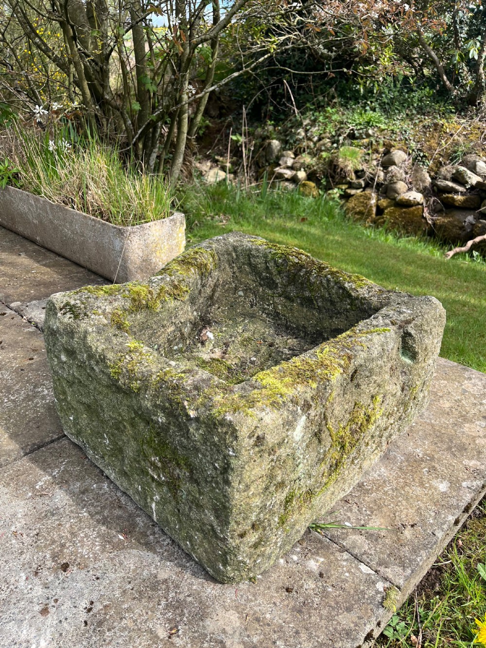 A rectangular hand-carved grey stone garden trough with a moss-covered weathered surface and a hollowed-out central basin.