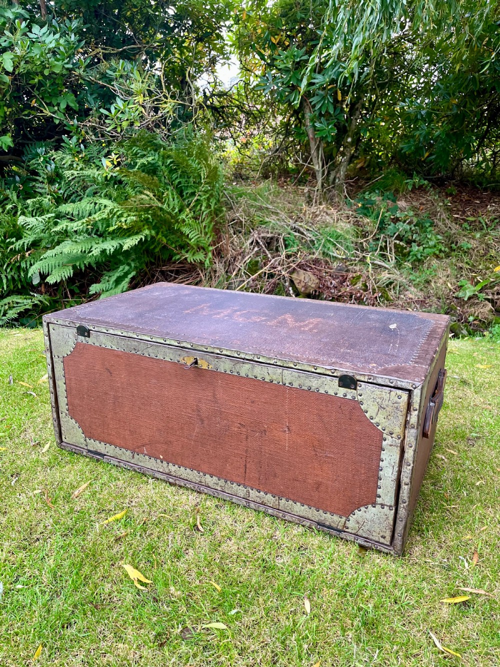 A rectangular antique dark brown canvas travel trunk featuring metal corner brackets, studding along the edges, and stencilled initials on top.