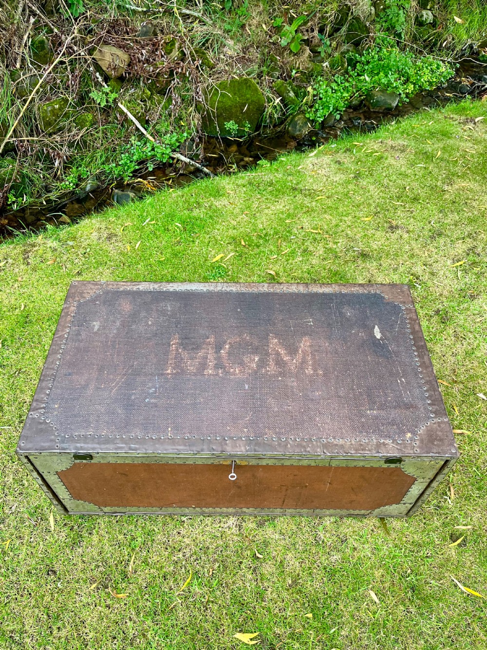 A rectangular antique dark brown canvas travel trunk featuring metal corner brackets, studding along the edges, and stencilled initials on top. View III