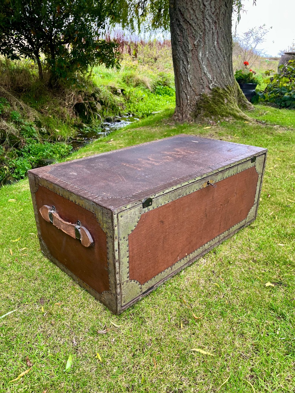 A rectangular antique dark brown canvas travel trunk featuring metal corner brackets, studding along the edges, and stencilled initials on top. View IV