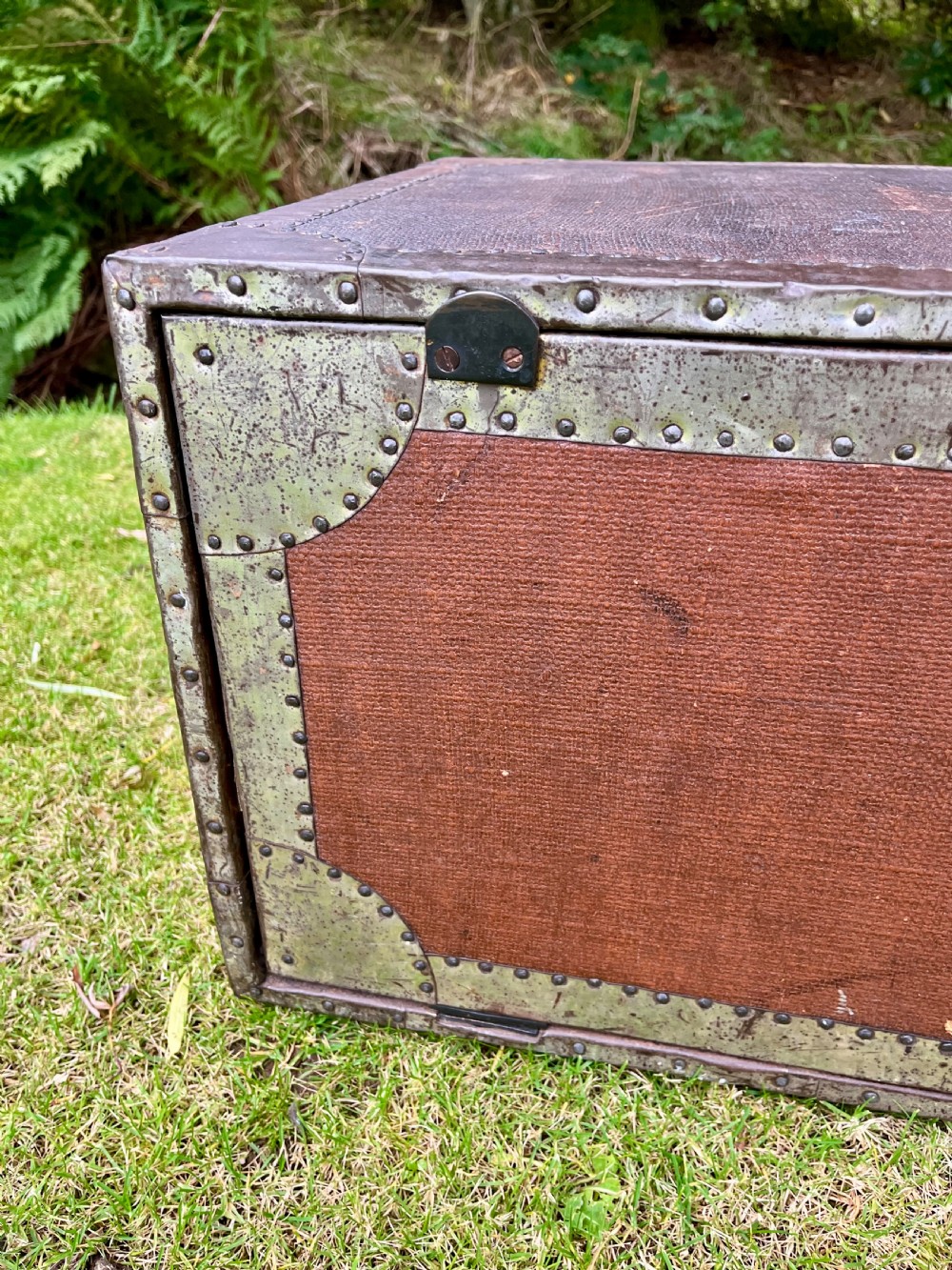 A rectangular antique dark brown canvas travel trunk featuring metal corner brackets, studding along the edges, and stencilled initials on top. View VI