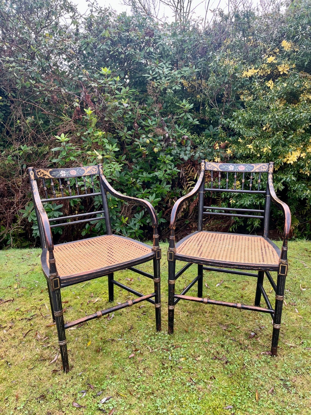 A pair of ebonised Regency armchairs featuring hand-painted floral cresting, turned spindle backs, swept arms, and woven cane seats. View II