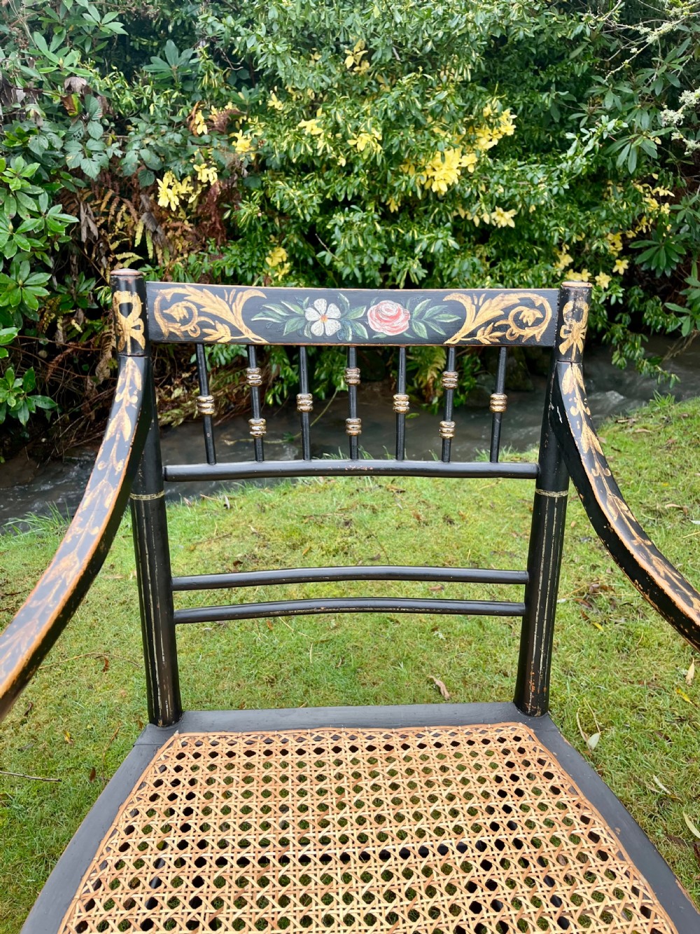 A pair of ebonised Regency armchairs featuring hand-painted floral cresting, turned spindle backs, swept arms, and woven cane seats. View IV