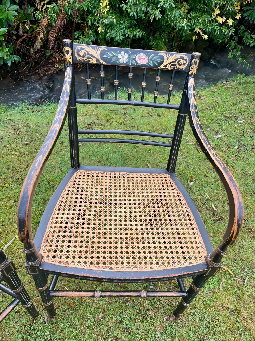 A pair of ebonised Regency armchairs featuring hand-painted floral cresting, turned spindle backs, swept arms, and woven cane seats. View V
