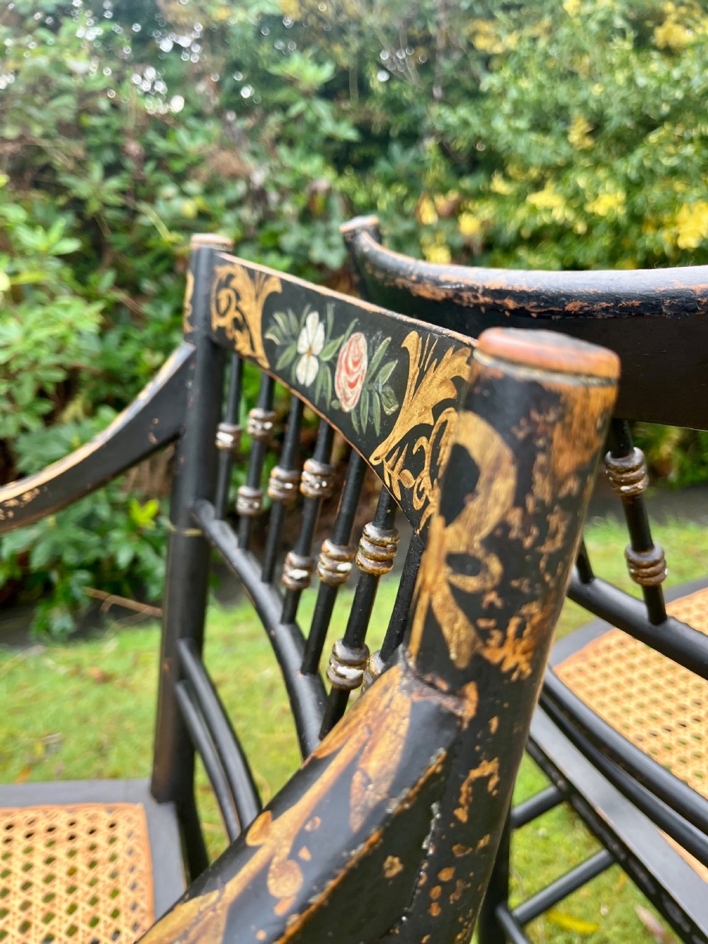 A pair of ebonised Regency armchairs featuring hand-painted floral cresting, turned spindle backs, swept arms, and woven cane seats. View IX