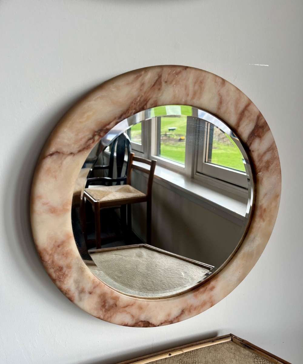 A circular wall mirror featuring a thick alabaster stone frame with visible pink and cream veining surrounding bevelled mirror glass.