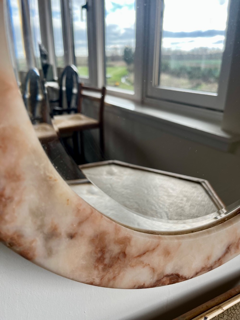 A circular wall mirror featuring a thick alabaster stone frame with visible pink and cream veining surrounding bevelled mirror glass. View III