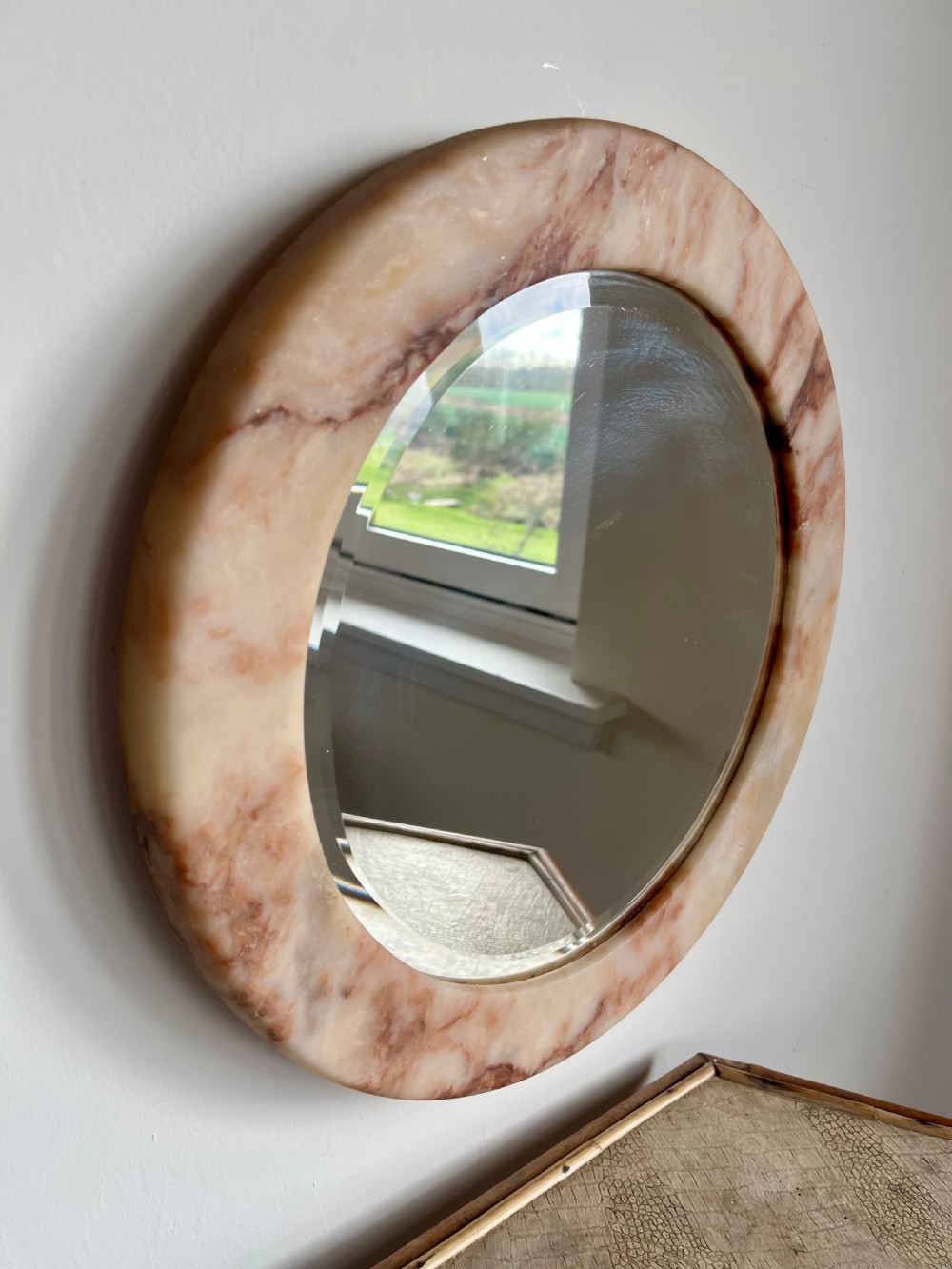 A circular wall mirror featuring a thick alabaster stone frame with visible pink and cream veining surrounding bevelled mirror glass. View IV