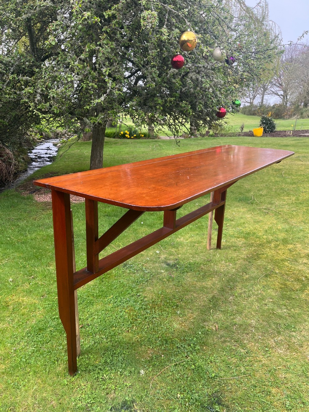 A long, narrow antique wooden butlers console table with a polished warm brown finish, rounded corners, and a collapsible trestle leg frame.