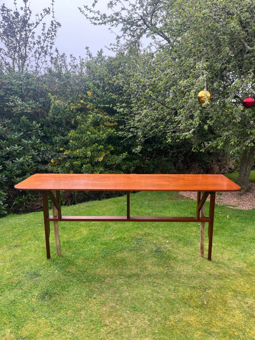 A long, narrow antique wooden butlers console table with a polished warm brown finish, rounded corners, and a collapsible trestle leg frame. View XI