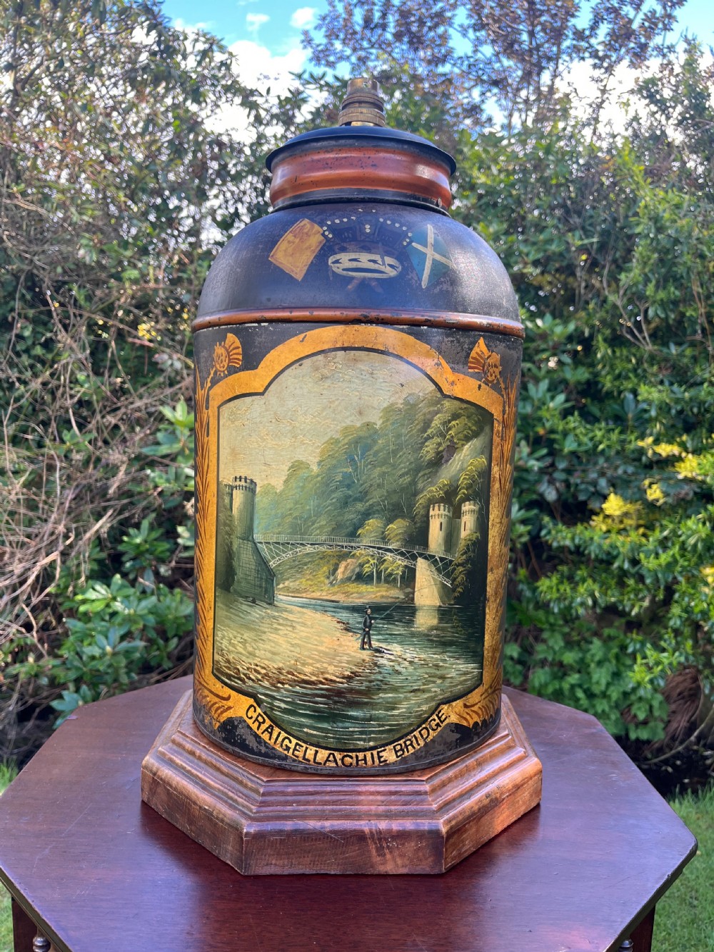 An antique dark metal tea canister converted into a table lamp, decorated with a hand-painted scene depicting Craigellachie Bridge set on a wooden base.