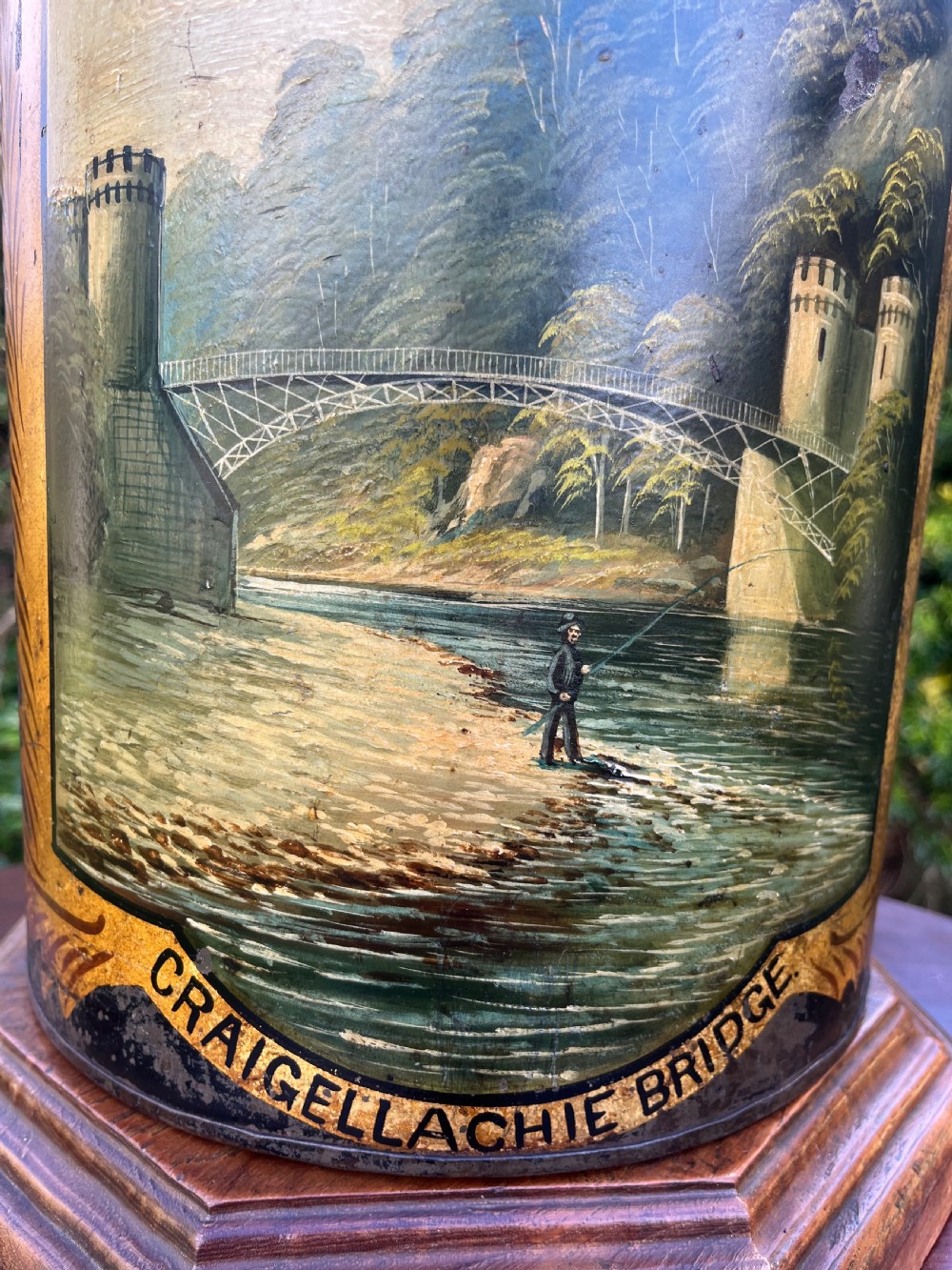 An antique dark metal tea canister converted into a table lamp, decorated with a hand-painted scene depicting Craigellachie Bridge set on a wooden base. View III