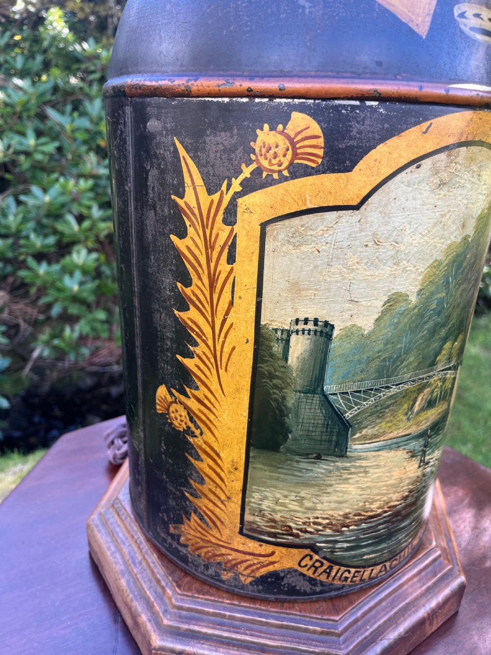 An antique dark metal tea canister converted into a table lamp, decorated with a hand-painted scene depicting Craigellachie Bridge set on a wooden base. View IV