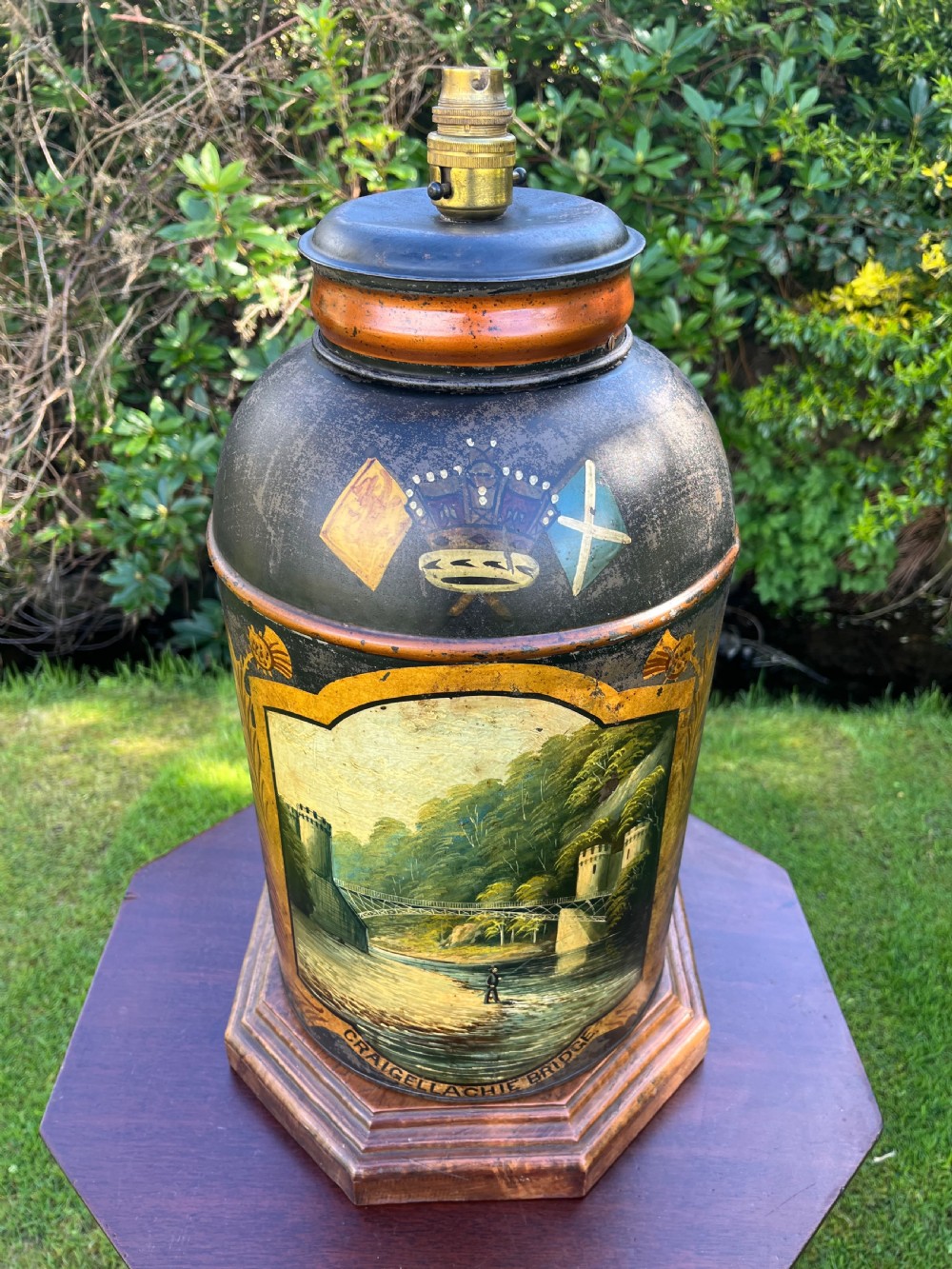An antique dark metal tea canister converted into a table lamp, decorated with a hand-painted scene depicting Craigellachie Bridge set on a wooden base. View VI