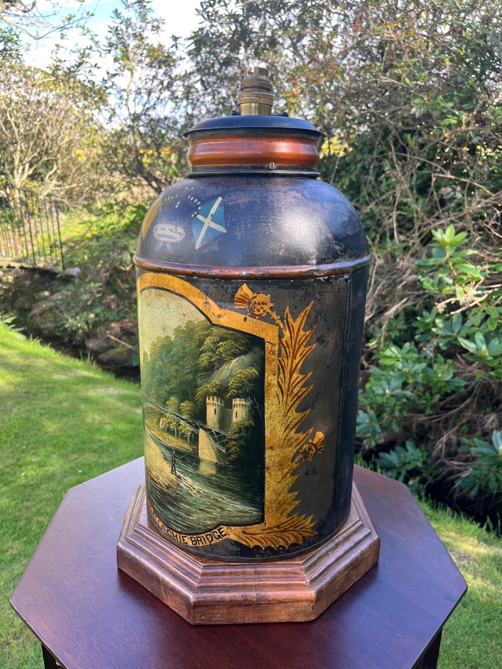 An antique dark metal tea canister converted into a table lamp, decorated with a hand-painted scene depicting Craigellachie Bridge set on a wooden base. View VII