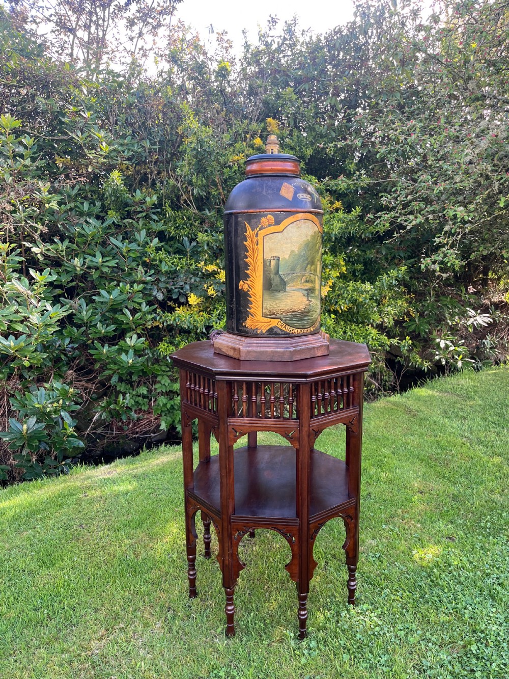 An antique dark metal tea canister converted into a table lamp, decorated with a hand-painted scene depicting Craigellachie Bridge set on a wooden base. View XI