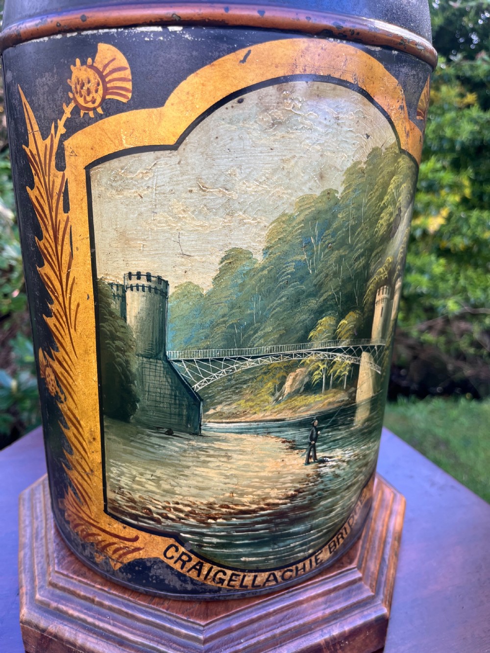An antique dark metal tea canister converted into a table lamp, decorated with a hand-painted scene depicting Craigellachie Bridge set on a wooden base. View XII