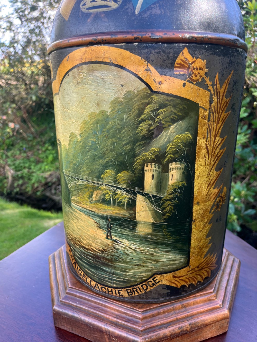 An antique dark metal tea canister converted into a table lamp, decorated with a hand-painted scene depicting Craigellachie Bridge set on a wooden base. View XIII