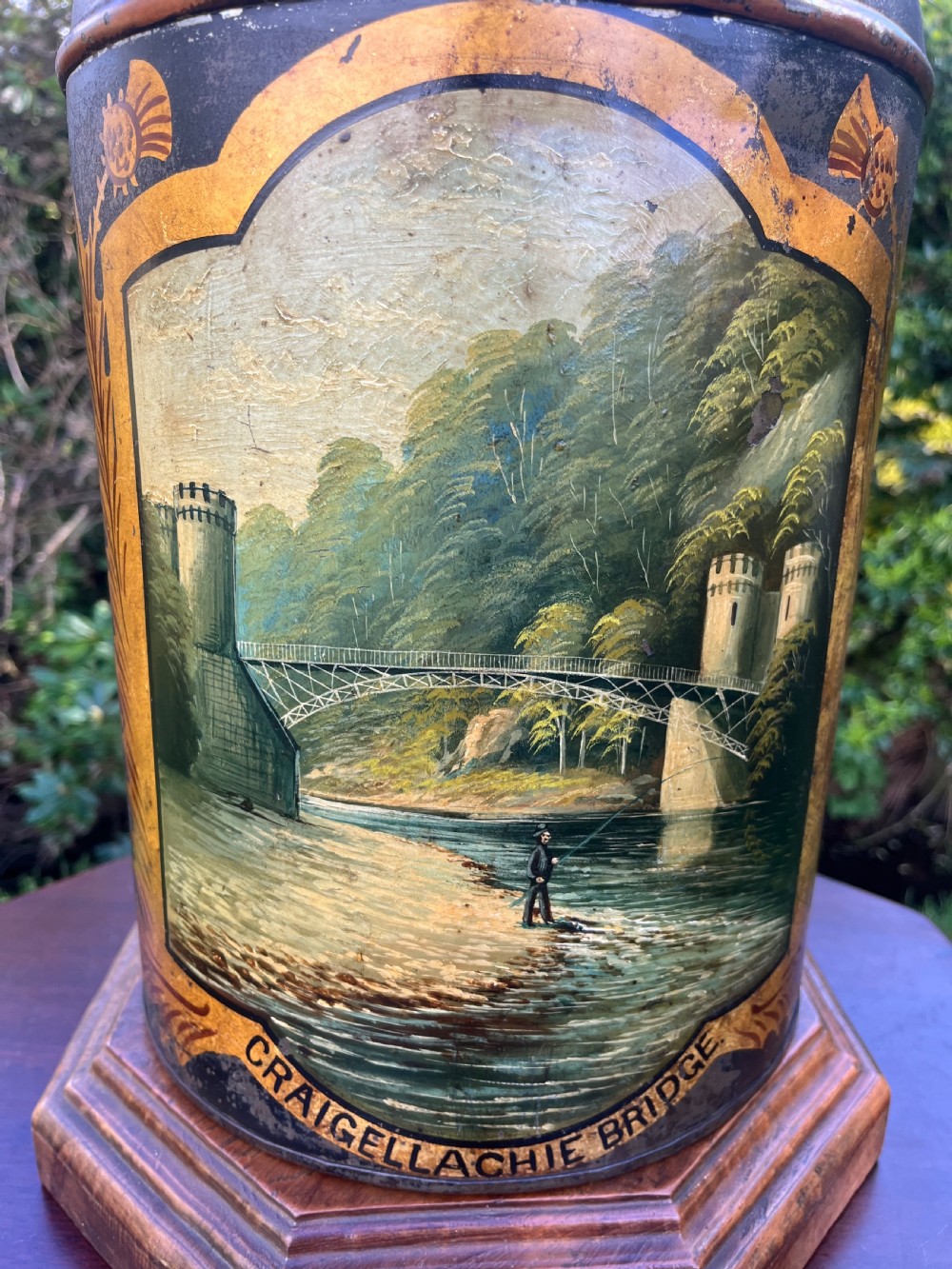 An antique dark metal tea canister converted into a table lamp, decorated with a hand-painted scene depicting Craigellachie Bridge set on a wooden base. View XV