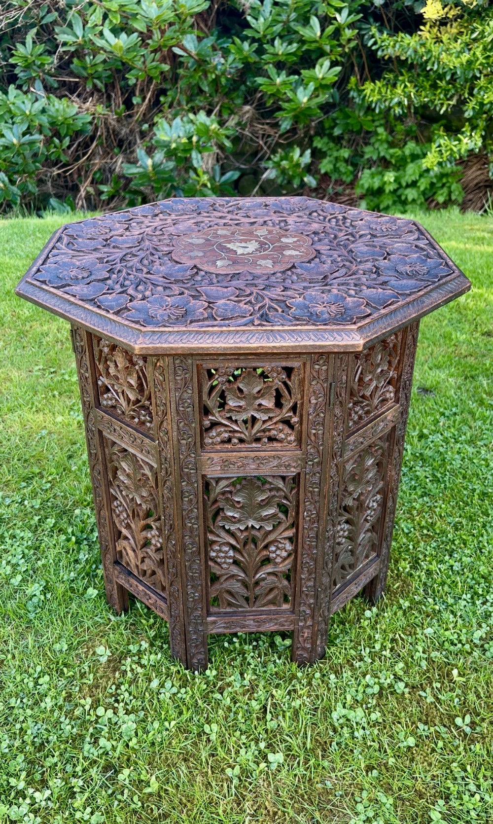 An octagonal dark wood occasional table featuring detailed foliate carvings and a central brass floral inlay on top, supported by carved openwork side panels.
