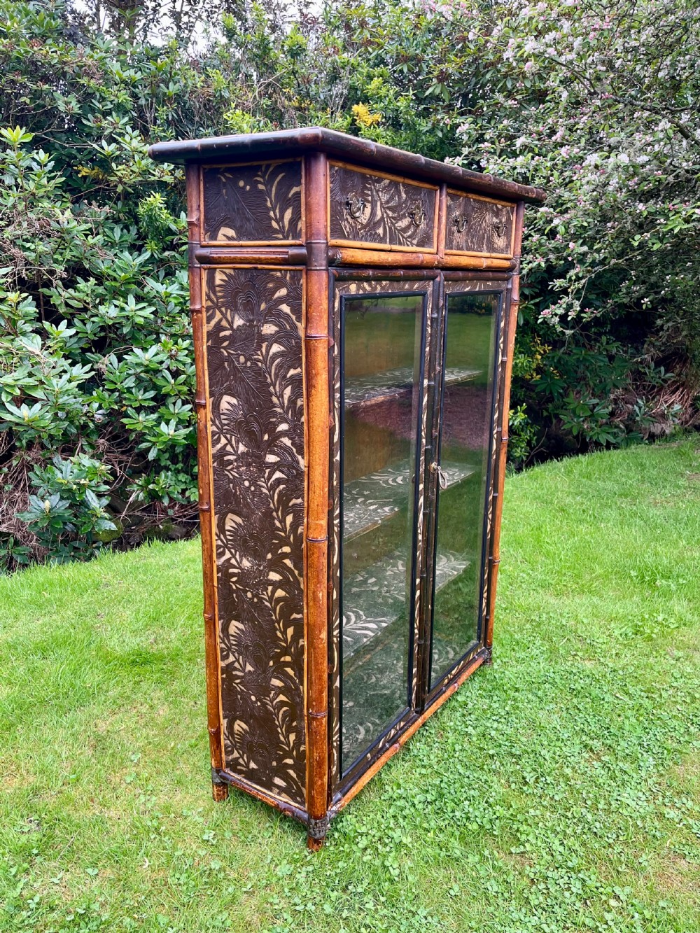 A late Victorian bamboo display cabinet with two glazed doors, upper drawers, and dark embossed botanical pattern panels.
