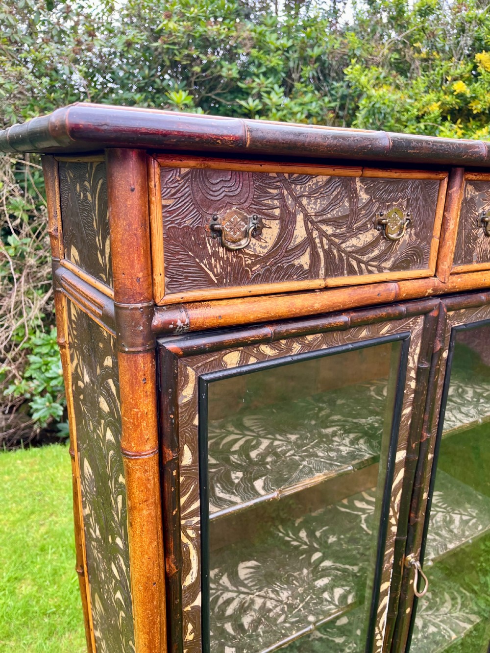 A late Victorian bamboo display cabinet with two glazed doors, upper drawers, and dark embossed botanical pattern panels. View III