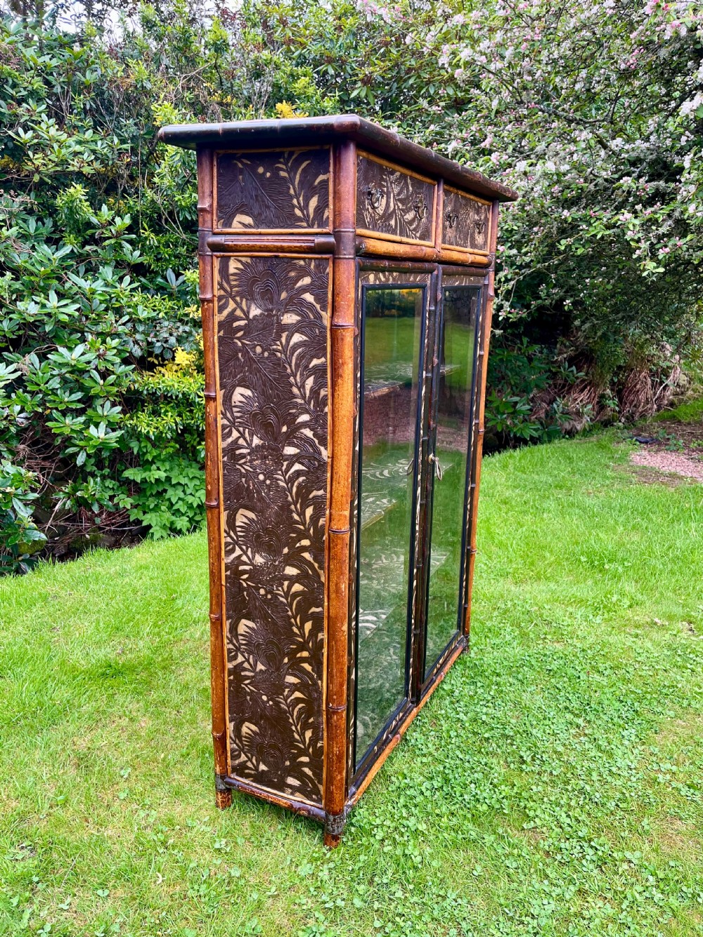 A late Victorian bamboo display cabinet with two glazed doors, upper drawers, and dark embossed botanical pattern panels. View XV
