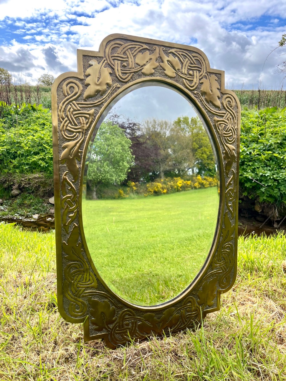 A large shaped brass wall mirror featuring an oval glass centre enclosed by relief Celtic knotwork and leaf motifs.