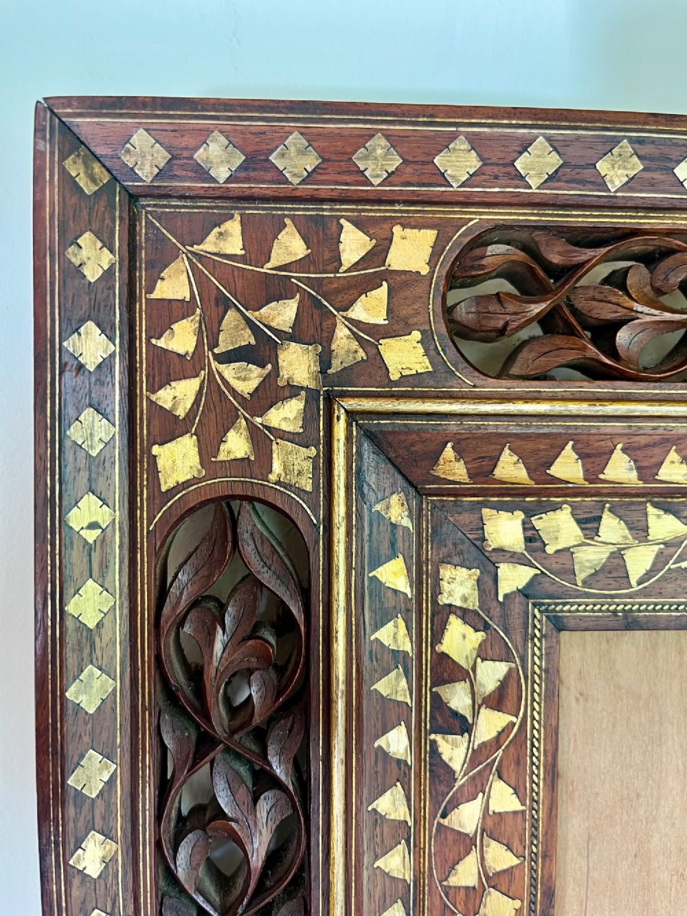 Corner detail of a rectangular dark wood frame decorated with brass foliate inlay and pierced scrollwork panels.