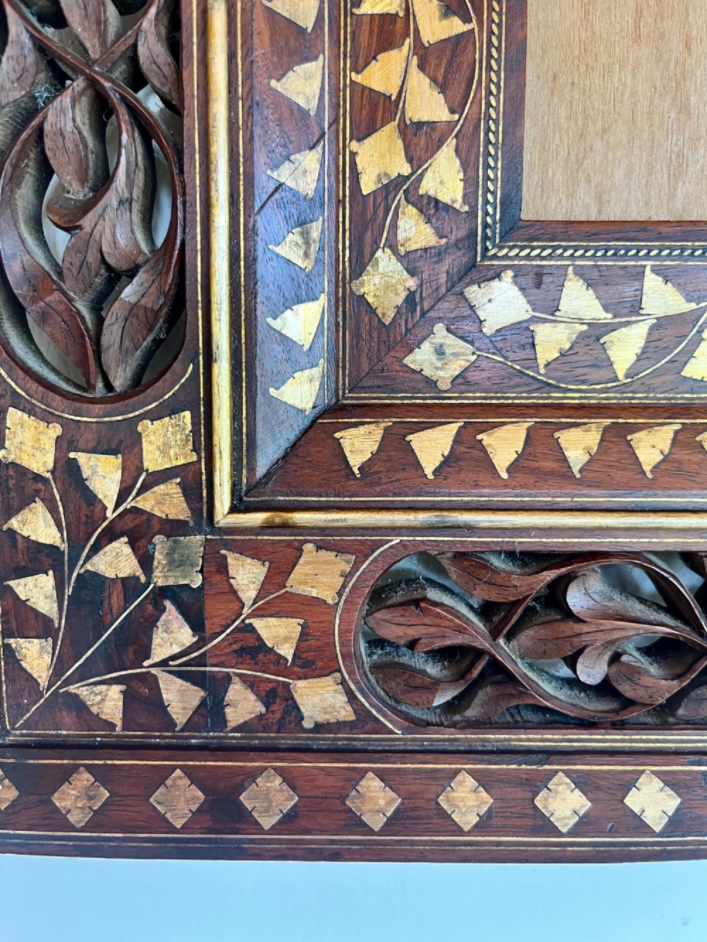 Corner detail of a rectangular dark wood frame decorated with brass foliate inlay and pierced scrollwork panels. View III