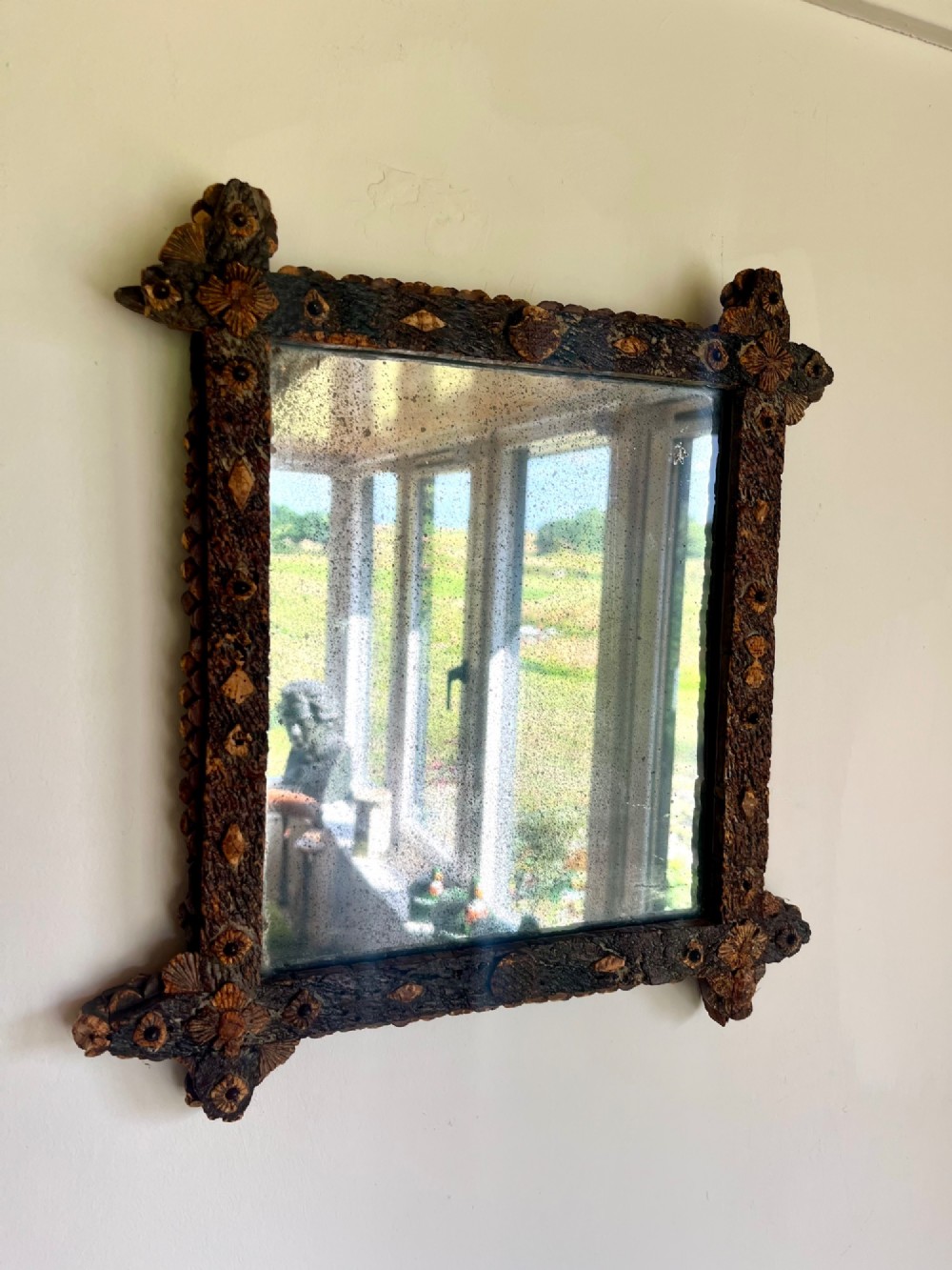 A square antique wall mirror framed with layered dark cork bark featuring intricate hand-carved rosettes and geometric notched corners surrounding foxed mirror glass.