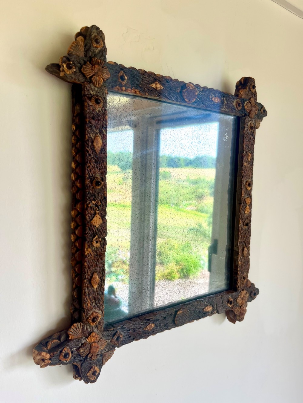 A square antique wall mirror framed with layered dark cork bark featuring intricate hand-carved rosettes and geometric notched corners surrounding foxed mirror glass. View IX