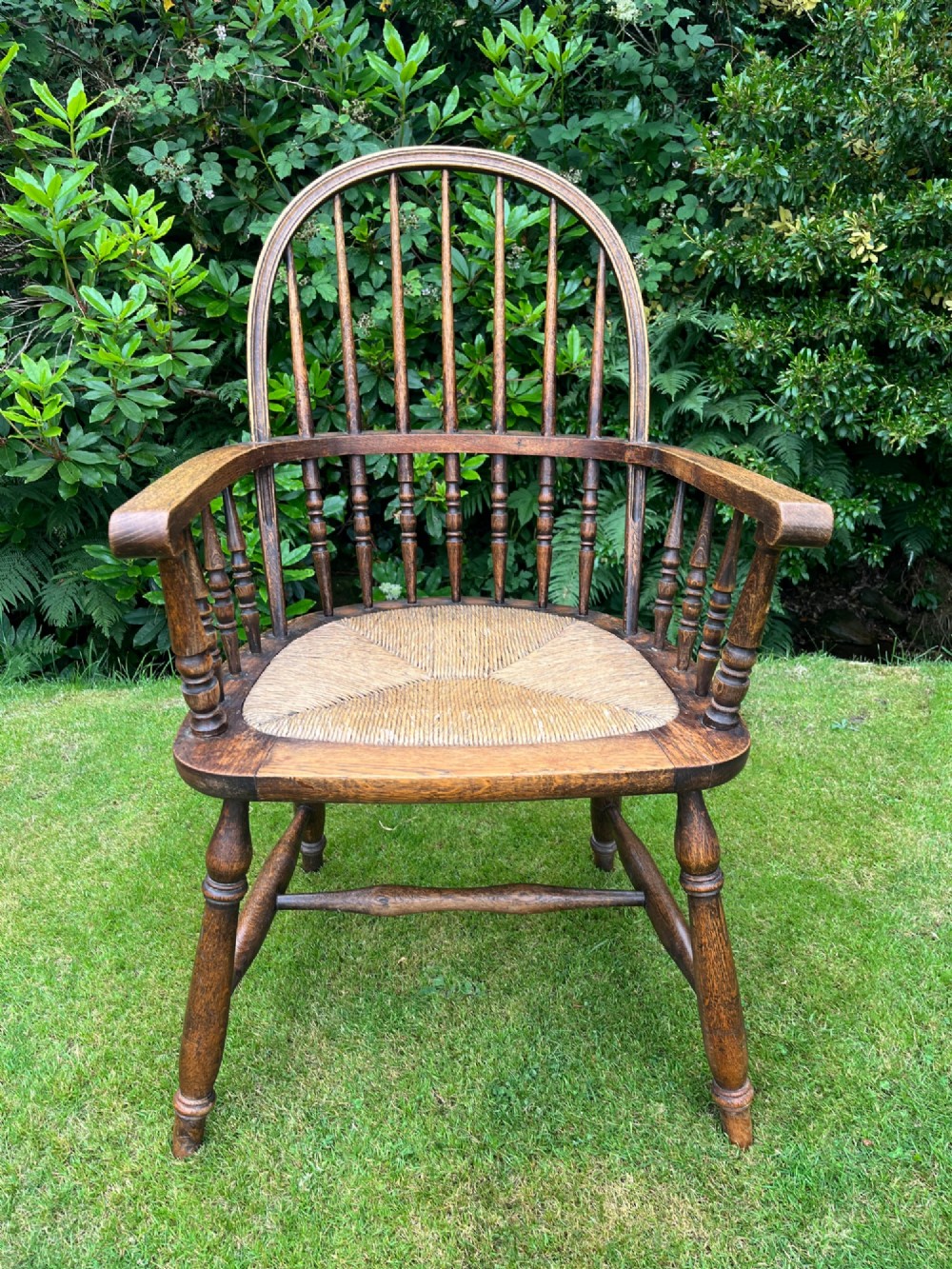 An antique ash Windsor armchair featuring a hoop back with vertical spindles, curved armrests, a woven rush seat, and turned legs linked by stretchers.