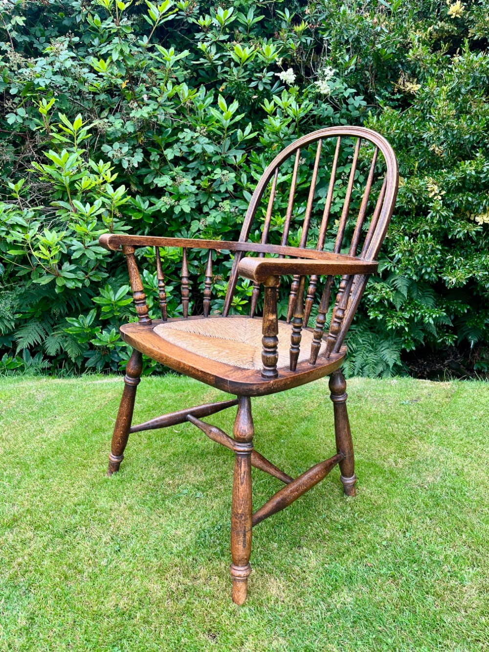 An antique ash Windsor armchair featuring a hoop back with vertical spindles, curved armrests, a woven rush seat, and turned legs linked by stretchers. View III