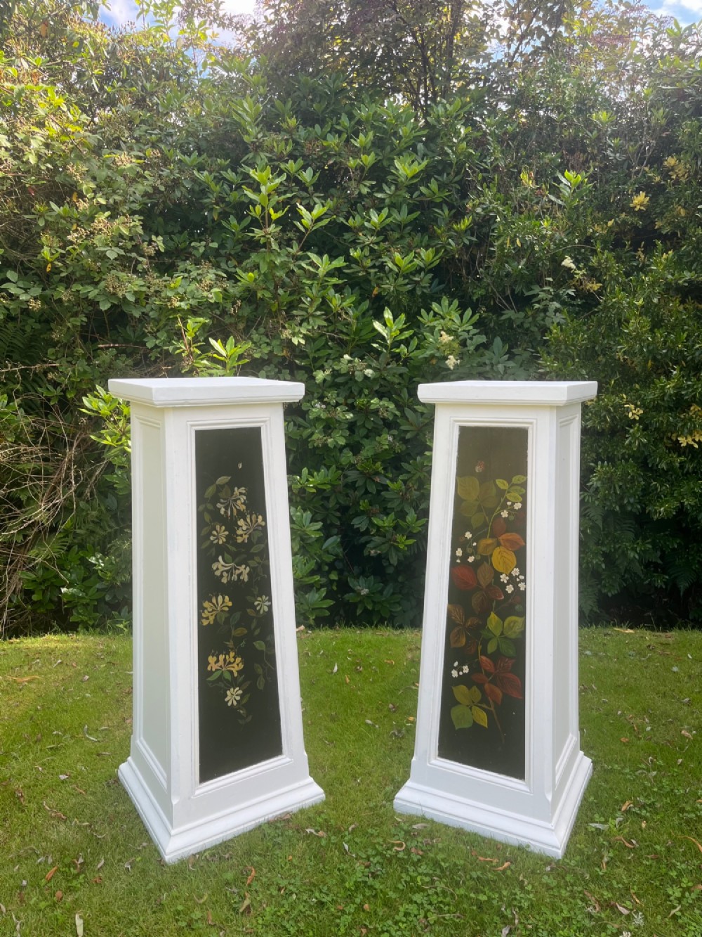 A pair of white wooden tapered display pedestals with inset black panels decorated with hand-painted floral and botanical foliage designs. View II