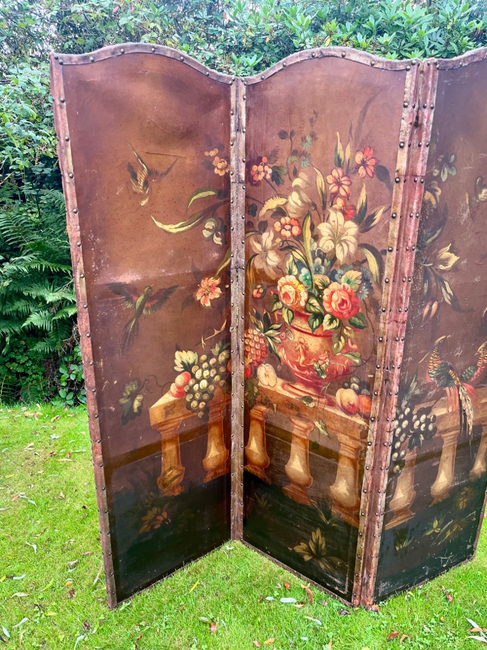 A three-panel antique folding leather screen hand-painted with a central urn of flowers, fruit, and exotic birds against a warm brown background. View II