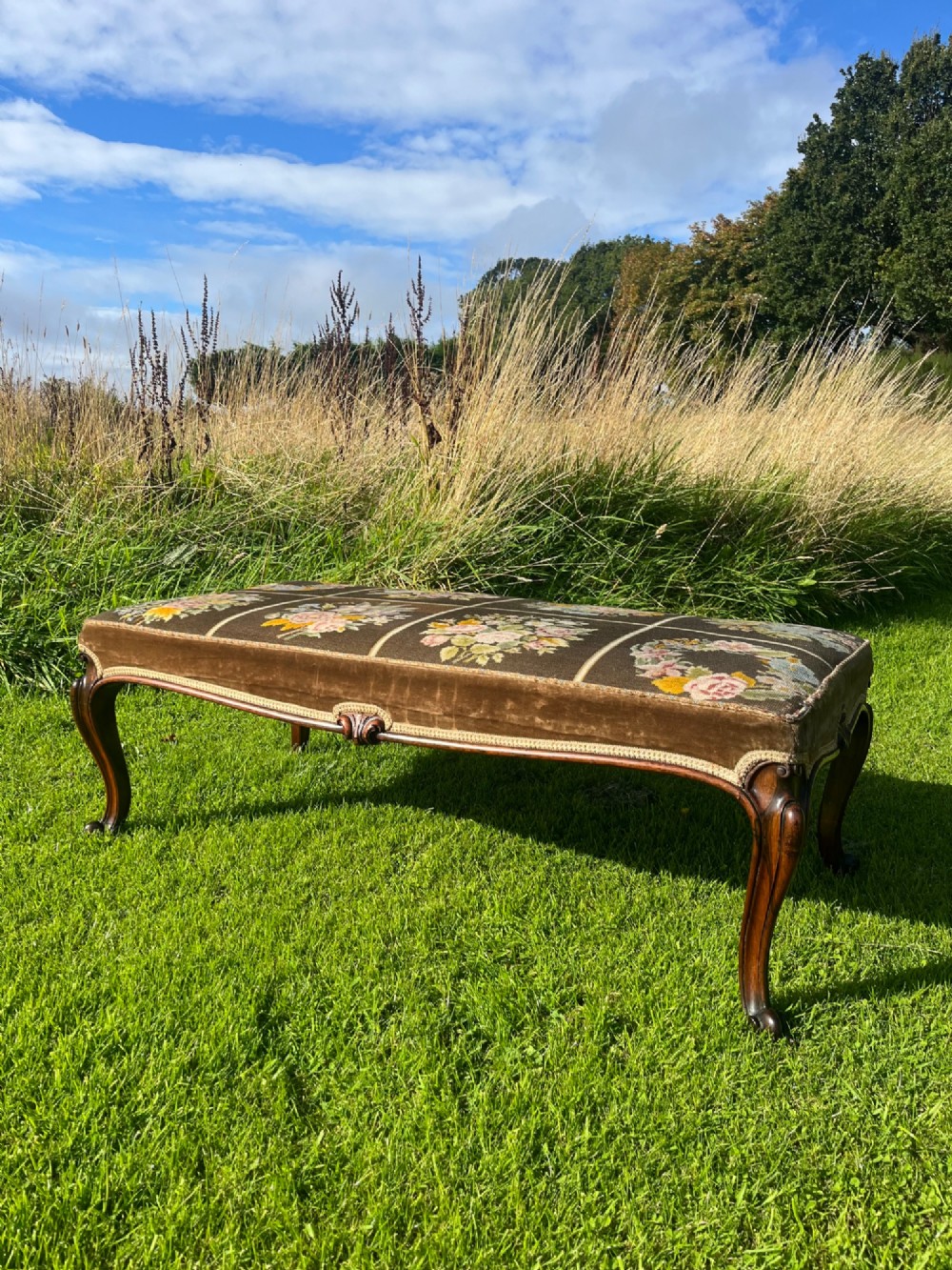 A long rectangular Victorian centre stool with dark rosewood cabriole legs and floral tapestry upholstery set in brown velvet.