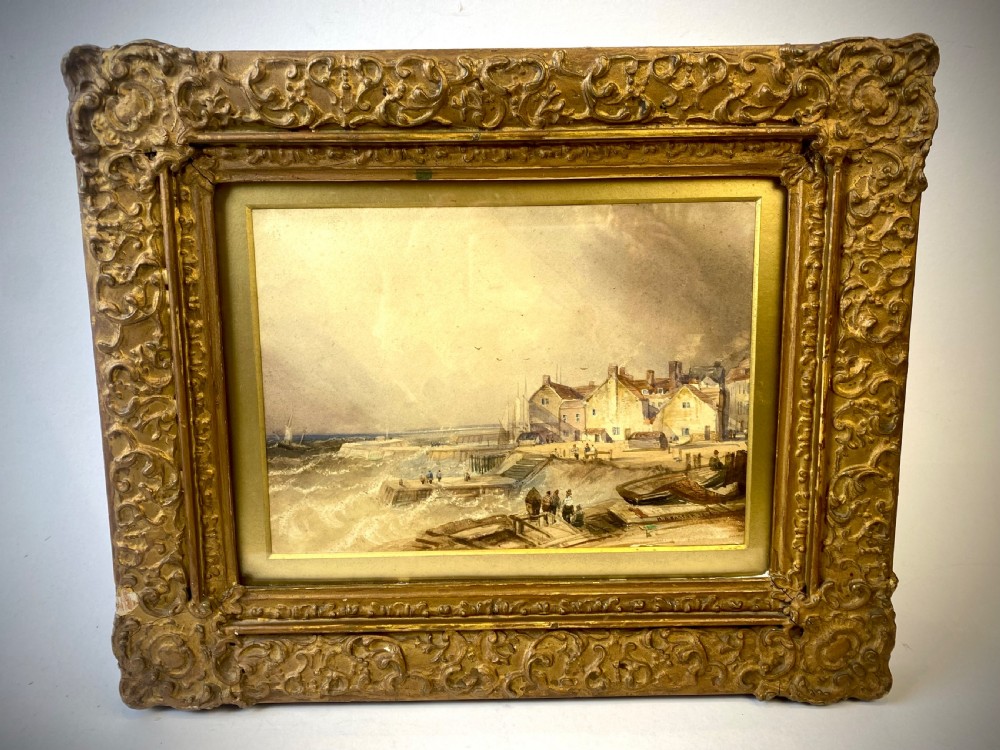 A 19th-century watercolour painting depicting a coastal town and harbour scene with rough seas, set inside a heavy gold gilt scrollwork frame.