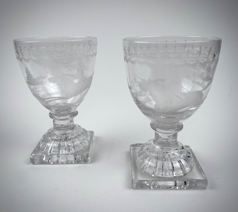 A pair of clear glass rummers featuring wheel-engraved fox hunting scenes set on square lemon squeezer bases.
