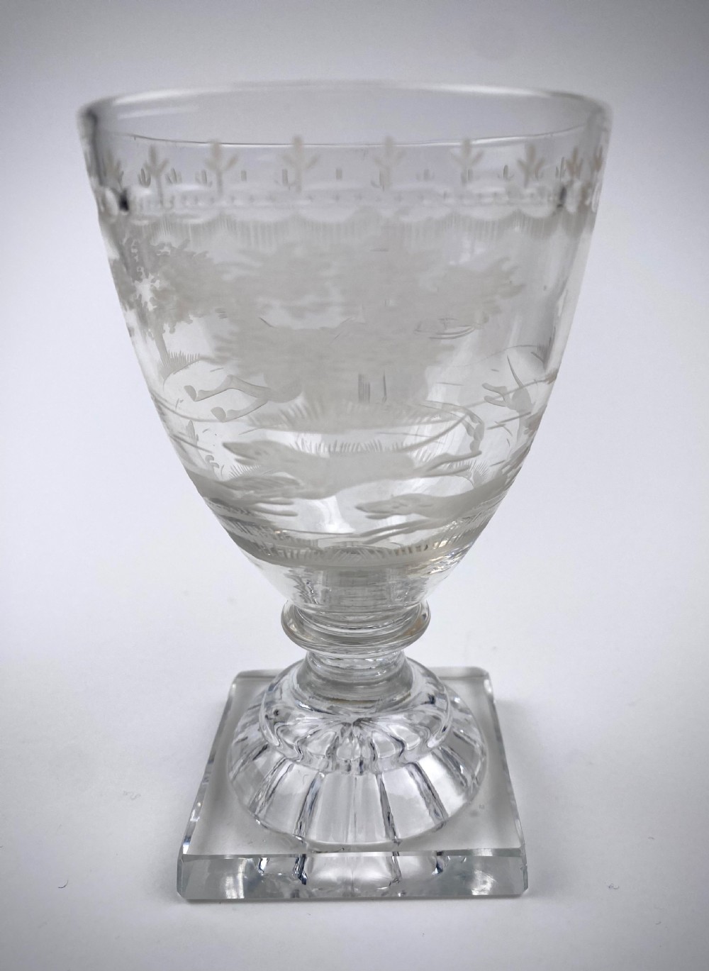 Clear glass rummer featuring an etched hunting scene of fox and hounds, supported by a ribbed stem and a square press-moulded lemon squeezer base.