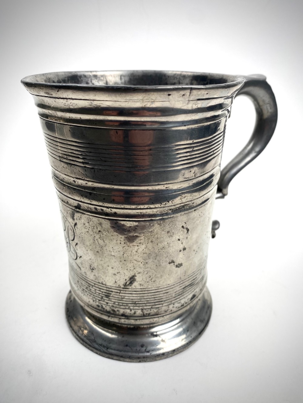An eighteenth-century Georgian pewter quart tankard with horizontal reeded bands, a cast handle, and an engraved monogram on its grey surface.