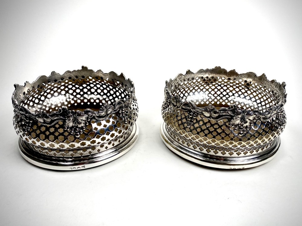 A pair of Georgian sterling silver wine coasters with pierced fretwork, grapevine rims, and dark wooden bases.